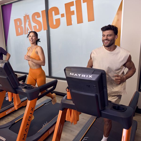 Een vrouw en een man rennen naast elkaar op loopbanden in een Basic-Fit club, terwijl ze glimlachen. Op de achtergrond zie je het grote Basic-Fit logo op het raam.